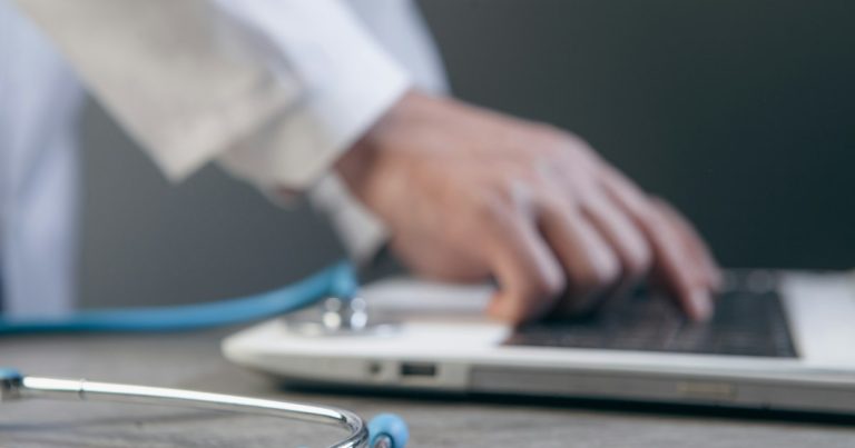 Doctor typing on a laptop with a stethoscope nearby, illustrating telemedicine for prisoners